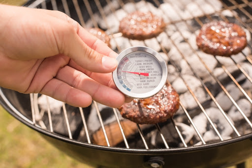 Photo of a hand holding a food thermometer, which is inserted in a hamburger on the grill.