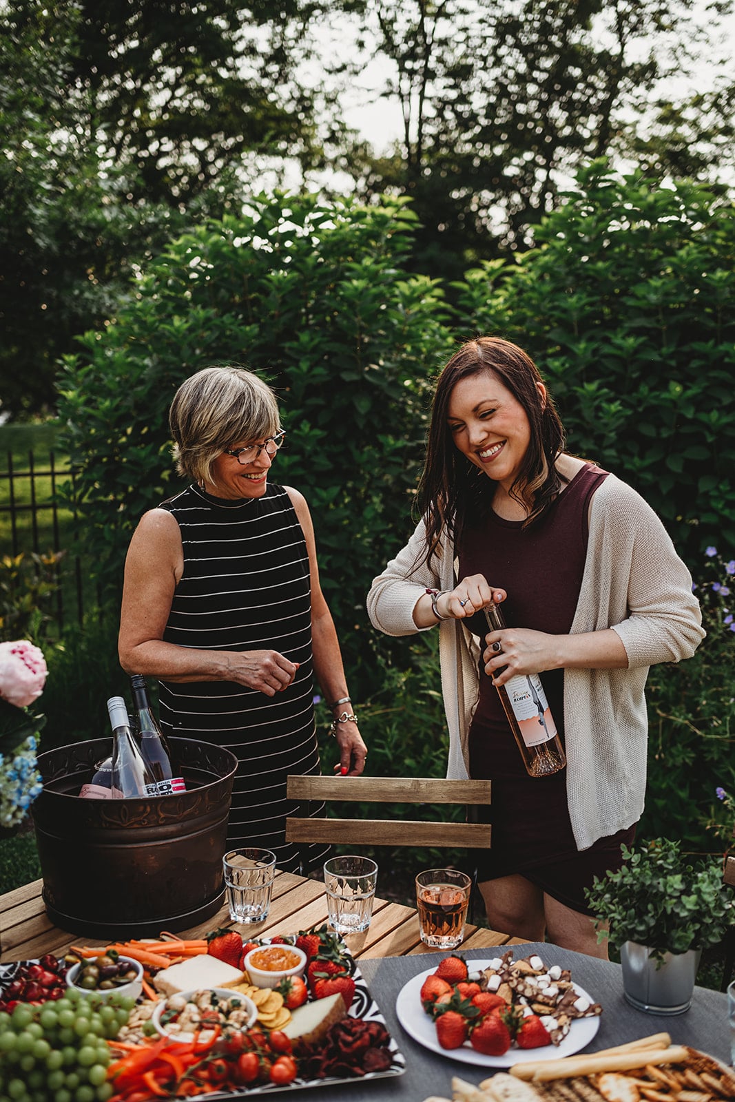 A photo of two women opening a bottle of wine at an outdoor gathering.