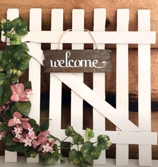 A photo featuring a wooden "Welcome" sign hanging on a white picket fence that is adorned with flowers.