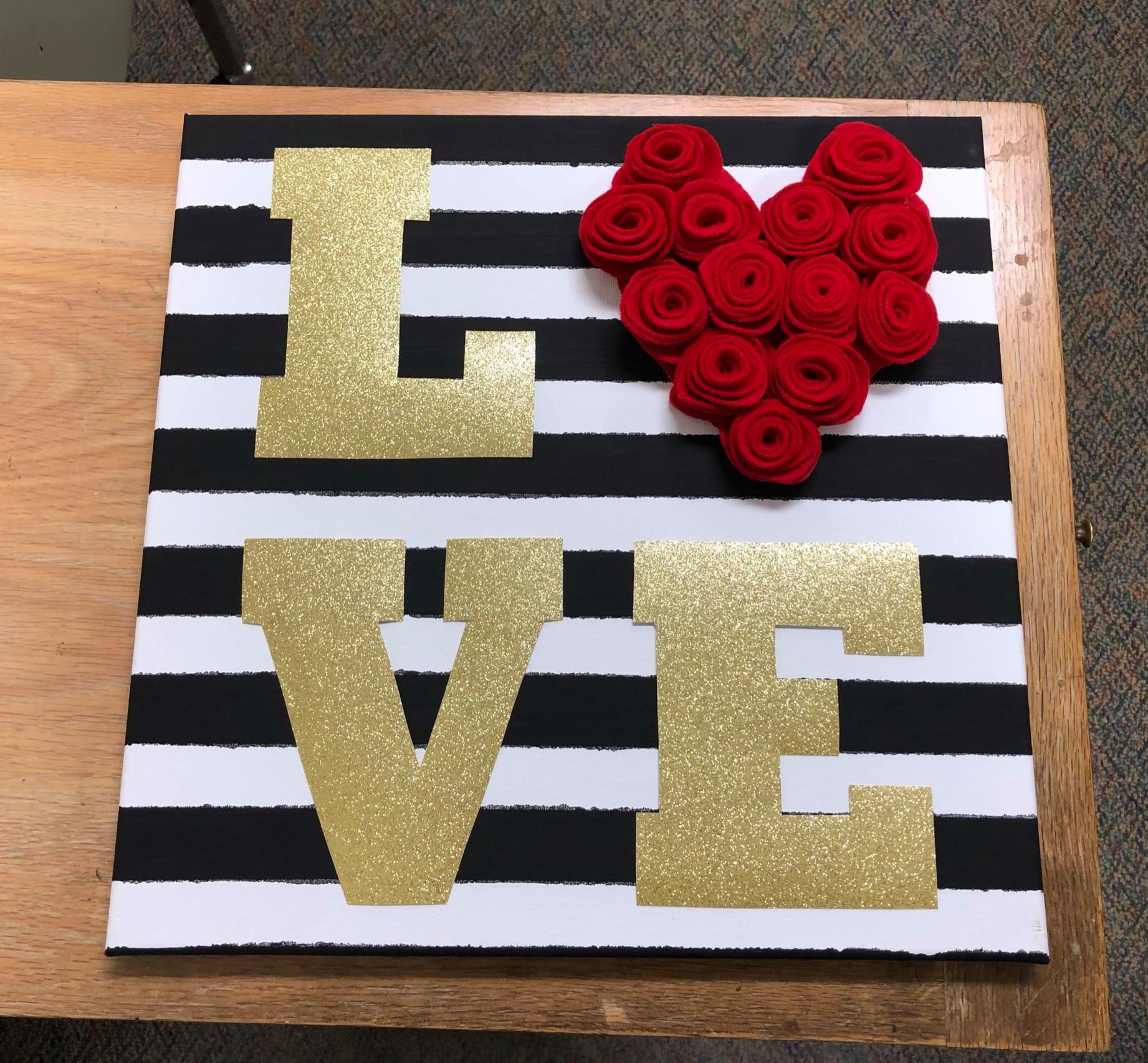 A photo of wall decor, featuring a square covered in black and white horizontal stripes. on top of that are the letters L, V and E on gold, arranged in a square. The O is a red heart.