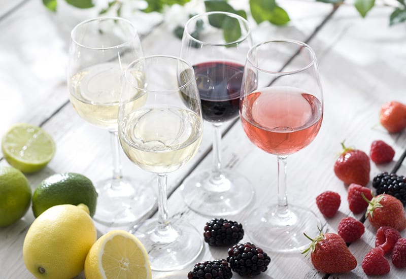 A color photo of four glasses of wine on a white wooden table surrounded by lemons, limes, and berries.