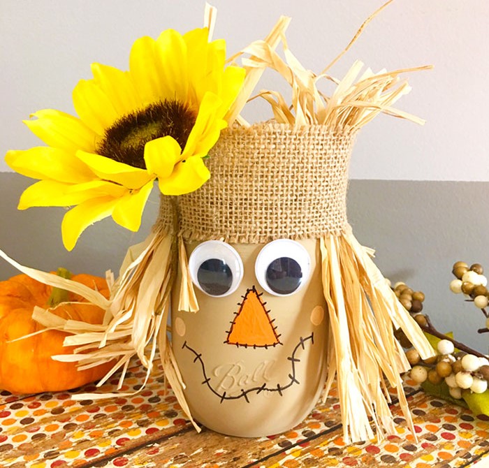 A photo of a scarecrow's head made from a mason jar, with a burlap hat, straw hair and a sunflower in its hat.