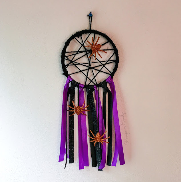 Photo of a spider web walling hanging featuring a hoop entwined with yarn to look like a spider web and purple and black streamers on the bottom.