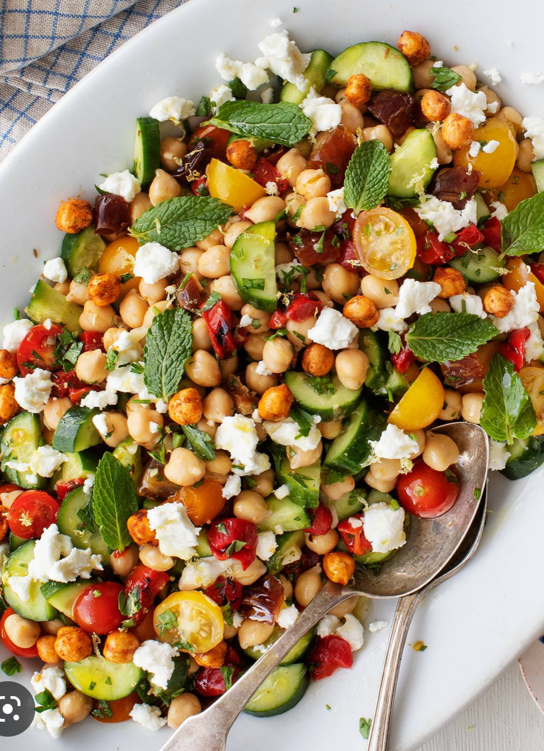 A color photo of a white platter filled with a salad made from chickpeas, cucumber, tomato, feta cheese and mint.