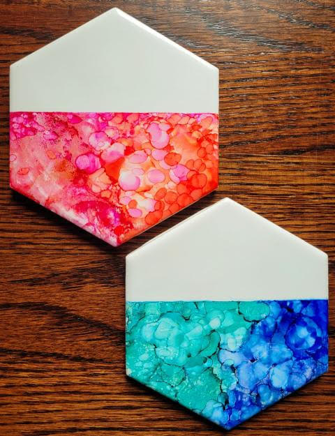 A color photo of two white hexagonal tiles, each half covered with an alcohol ink design. One is in the red color family, the other blues and greens.