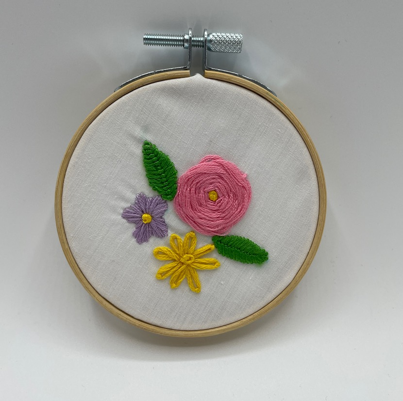 A color photo of white fabric in an embroidery hoop with three flowers stitched onto it, in yellow, pink and purple thread..