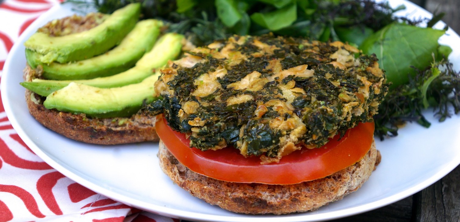 A color photo of a kale burger sitting atop a slice of tomato and the bottom of a bun, with the top of the bun an avocado slices alongside.
