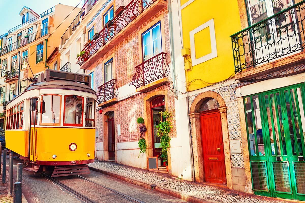 A color photo of a trolley car passing in front of colorful buildings on a narrow street in Lisbon, Portugal.