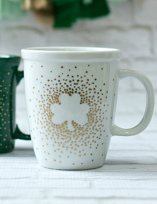 A photo of a white mug adorned with a negative space shamrock shape created by outlining it with dots of gold paint.it
