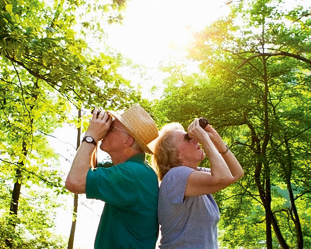 An older couple in the woods standing back to back each looking through binoculars.