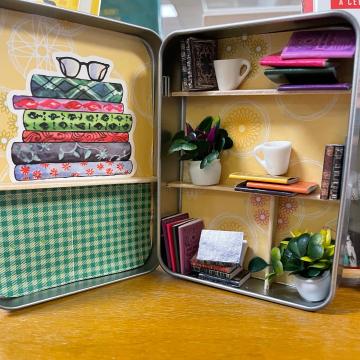 A color photo of a mini library, featuring wallpaper, shelves and tiny books, plants and trinkets.