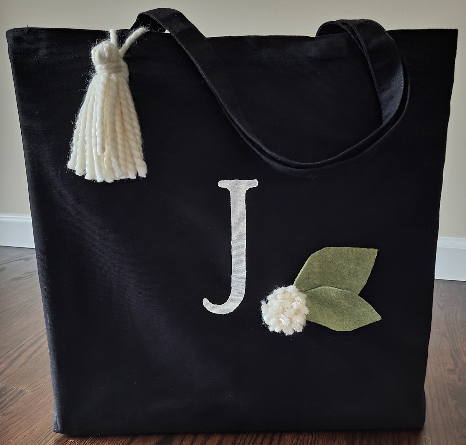 A photo of a black tote bag decorated with the letter J, a leaf applique and a tassel.