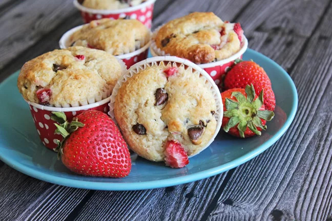 A color photo of four strawberry chocolate chip muffins and two fresh strawberries on a blue plate.