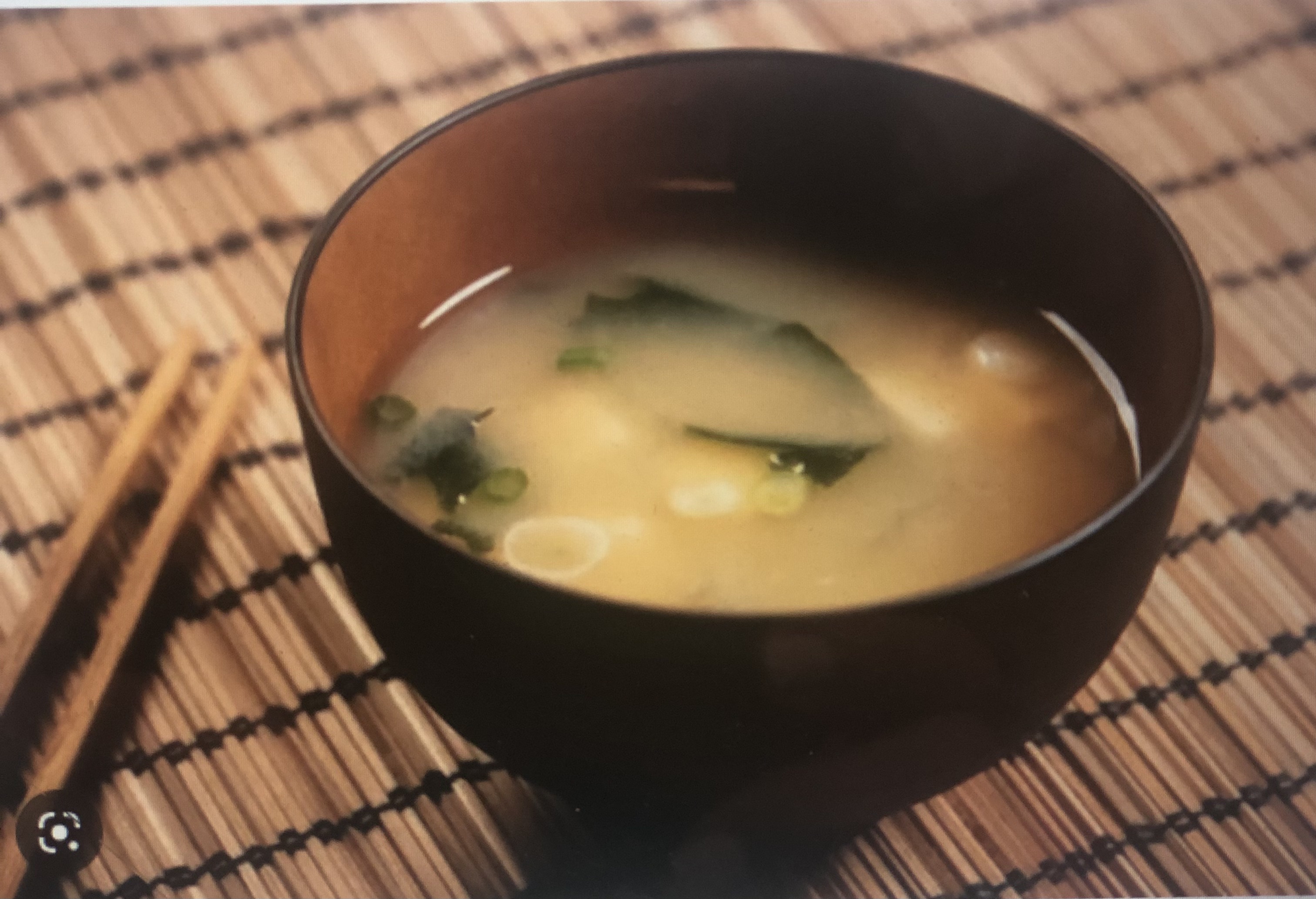 A color photo of a bowl of miso soup on a bamboo placemat, a pair of chopsticks laying beside..
