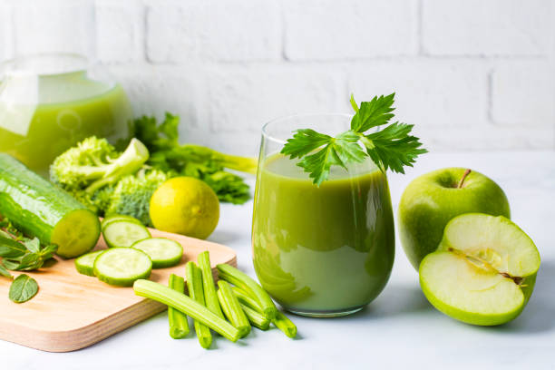 A color photo of a glass filled with green juice surrounded by ingredients such as avocado and celery.
