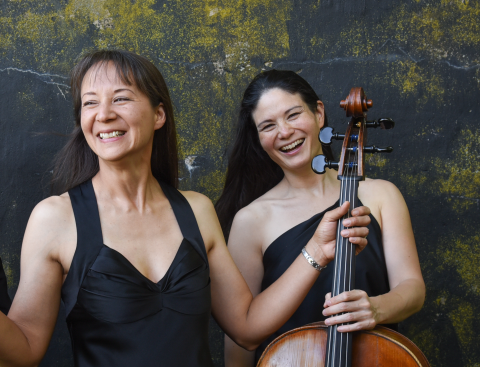 a color photo of pianist Naomi Niskala on the left and cellist an-Lin Bardin on the right. They are both holding a cello and laughing.