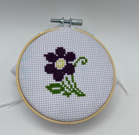 A color photo of a purple flower cross stitched on white fabric, which is stretched into a embroidery hoop.