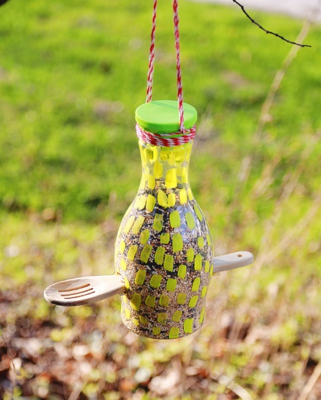 A color photo of a bird feeder made from a plastic juice bottle and featuring a wooden spoon as a perch.
