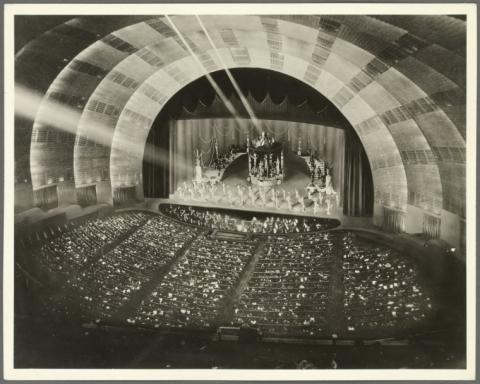 A sepia toned photo of the inside of Radio City Music Hall featuring the stage, seating and the soaring interior.