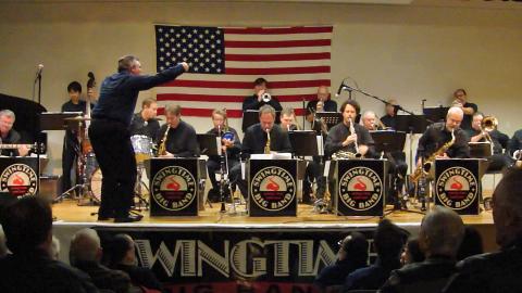 A color photo of the Swingtime Band performing on stage at the library with an American flag in the background.