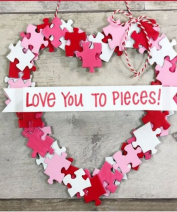 A heart-shaped wreath made from white, pink and red puzzle pieces and adorned with a banner that says, Love You to Pieces.