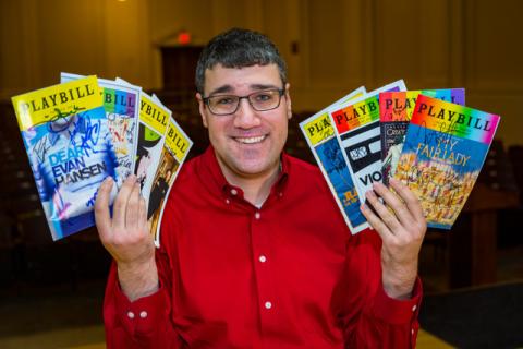 A color photo of Brian Stoll holding up several issues of Playbill in each hand.