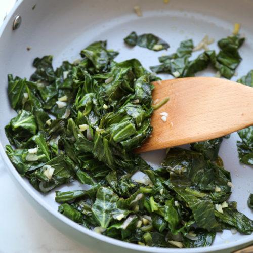 A photo of greens sauteing with a wooden spatula moving them around the pan.