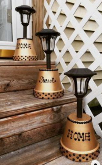 A color photo of three upside down flower pots fitted with solar lanterns and sitting on three separate steps. The pots are painted gold and feature the words Home, Sweet and Home.