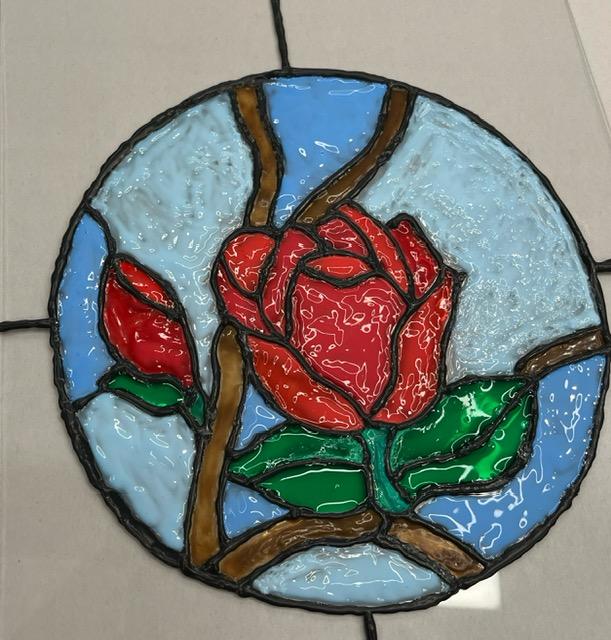 A color image of a round piece if art featuring a red rose on a blue background that looks like stained glass.