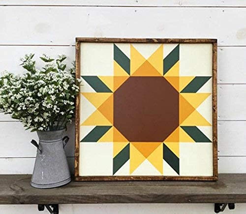 A color photo of a framed sunflower quilt square sitting on a shelf next to a small vase of flowers.
