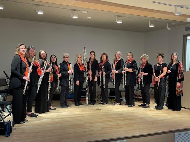 A photo of the members of the Long Island Flute Club Choir holding their flutes and standing in a semi-circle on a stage.