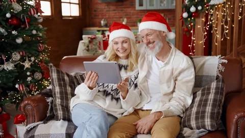 A couple dressed in Santa hats video chatting with someone on a tablet.